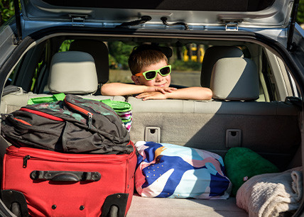 Enfant portant des lunettes de soleil appuyé à l'arrière d'une voiture chargée de valises et sacs pour les vacances d'été